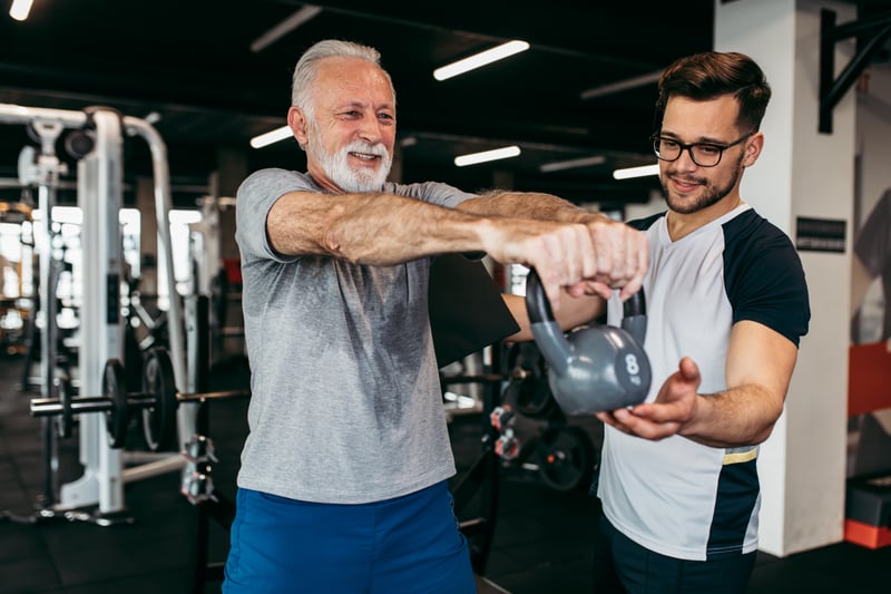 Man with a white beard lifting a kettle bell under the guidance of a personal trainer